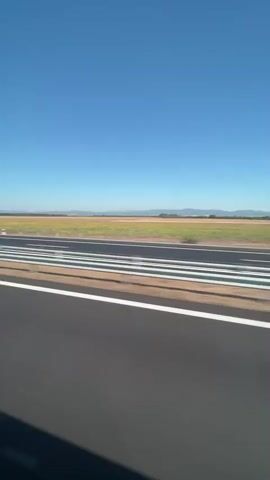 The video shows a view from a moving vehicle, looking out at an airport runway. The sky is a clear blue. The runway is paved and has multiple white lines painted across it, suggesting it is a taxiway or part of the runway itself. Beyond the runway, there are fields of brown and green vegetation, and in the distance, there are rolling hills or mountains. The movement of the vehicle causes a slight blur, particularly in the foreground where the road surface is visible. The shadow of the vehicle or its occupants can be seen in the bottom right corner of the frame.