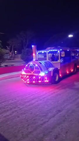 At night, a light-up, faux train makes its way across a cobblestone parking lot. The locomotive-style lead car is decorated with red, blue, and white lights, and sounds an air-raid siren-like horn.  Two passenger cars are pulled behind, each with a partial canopy.  People are visible in the passenger cars.  Street lamps and trees stand in the background.