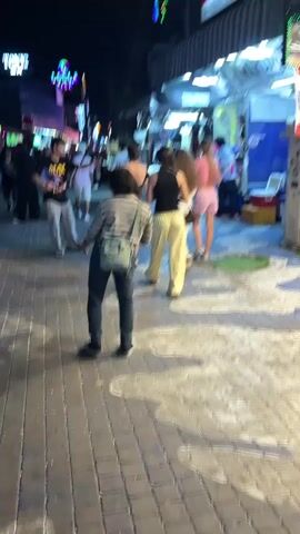A low-angle, handheld video shows a paved walkway at night, lined with shops. In the foreground, a person wearing a striped shirt, dark pants, and a light green crossbody bag dances with their back to the camera. The walkway is illuminated by overhead lights and neon signs from the shops. In the mid-ground and background, several other people are walking and standing. One shop appears to be selling clothing.