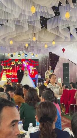 The video captures an indoor wedding ceremony or reception in Bình An, Vietnam, with a large gathering of guests. In the foreground, numerous people are seated at tables, watching the stage. A woman in a striped shirt is seen recording the event with her phone.

On the decorated stage, a bride and groom stand prominently. The bride wears a white gown and holds a floral bouquet, while the groom is in a black suit. Behind them is a large screen displaying red lanterns and the Vietnamese text "Lễ Tân Hôn" (Wedding Ceremony) along with the names "Quang Trọng" and "Ngọc Ý," and "Ngày 6." To the right of the couple, a person plays a keyboard. Another woman, possibly the master of ceremonies, is visible speaking into a microphone. The ceiling is elaborately decorated with white draped fabric and hanging yellow lantern-like lights, contributing to a festive atmosphere. Large speakers are positioned to the right of the stage. The guests appear attentive and engaged in the event.