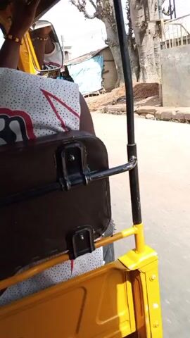 Here's a description of the video: 

The video appears to be shot from the back of a moving auto-rickshaw, also known as a tuk-tuk. The tuk-tuk has a yellow frame.  In the foreground, you can see the back of a passenger wearing a white shirt with a red and black pattern, and a brown seat. The view shows the streets of what looks like a bustling town or city with buildings and shops on either side. Other vehicles, including a blue truck, can be seen passing by. The sky is bright and sunny.