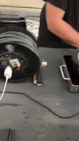 Here's a description of the video:

The video begins with a close-up of a power strip reel connected to a phone charger, which is charging a phone. The camera shifts to a man placing a frying pan full of what appears to be cooked dumplings or similar food items onto a portable induction cooktop. The cooktop's surface glows pink, indicating it is active. Finally, the camera briefly shows the lower legs of two people. One person is wearing beige Crocs, and the other is wearing darker footwear with olive green pants. The setting appears to be in a workshop or garage type environment due to the presence of a metal work surface and concrete floor.