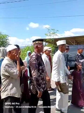 The video shows a group of people, predominantly men wearing traditional Islamic attire including peci hats, walking in a procession. Some women in hijabs are also visible, some appearing to be in prayer or observing. The setting appears to be outdoors, possibly a street or pathway, with lush green trees and hills in the background under a clear blue sky. A vehicle is parked on the right side of the frame. The timestamp indicates the video was recorded on June 25, 2026, at 10:44 AM in the Bireuen Regency.