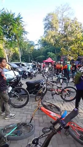 A crowd of cyclists and spectators gather outdoors. Several bicycles are parked in the foreground, with some lying on the ground. In the background, there are many people, motorcycles, cars, and tents. The scene appears to be an outdoor event or gathering, possibly a cycling race or rally, in a paved area surrounded by trees. The lighting suggests it is daytime, likely morning or late afternoon. Some individuals are wearing cycling gear, including helmets.