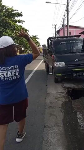 A person wearing a blue t-shirt with the word STAFF on it in white letters and a white baseball cap is walking on the side of a paved road. The person is also wearing burgundy shorts and white sneakers with black accents. The person is walking away from the camera and is on the left side of the road. To the right of the person, parked on the side of the road, is a black pickup truck. The truck appears to be partially stuck in a hole or ditch next to the road. The ditch is uneven and appears to have some debris in it. The road appears to be in an urban or semi-urban area, with trees and buildings visible in the background. The sky is overcast.