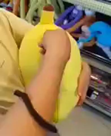 A person's hands are visible, holding a yellow plush banana. The person unzips the banana, revealing a plush monkey inside. The person then pulls the monkey out of the banana peel. The background is blurred, but appears to be a retail environment with shelves of merchandise.