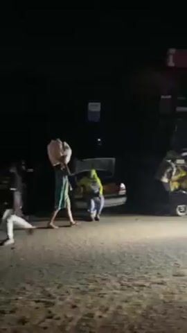 At night, several people are loading a car parked on the side of the road. The car's trunk is open, and individuals are filling a wheelbarrow with items from the trunk. One person stands nearby with a bag on their head, while another car passes in the background. A sign is visible in the background as well.