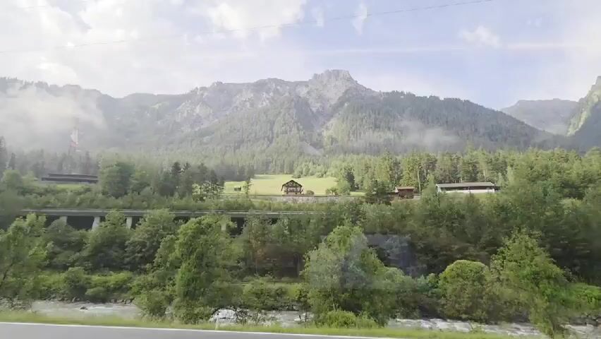This media file captures a scenic view of the Austrian Alps near Flirsch. The scene is dominated by a majestic mountain range in the background, partially obscured by soft clouds and mist, suggesting a slightly overcast or humid day, possibly in the morning or late afternoon. Dense green forests cover the lower slopes of the mountains, with a scattering of trees in the foreground.

A prominent feature in the mid-ground is an elevated, possibly abandoned, structure resembling a viaduct or an old railway line, supported by numerous concrete pillars and arches, now overgrown with vegetation. Above this structure, nestled in a clearing on a hillside, are a few buildings, including a traditional-looking house with a dark roof and wooden elements, hinting at human habitation in this otherwise wild landscape. A tall, lattice-style electricity pylon stands to the left of the frame, indicating the presence of modern infrastructure amidst the natural beauty.

In the foreground, a fast-flowing river or stream runs parallel to a paved road, suggesting a transportation route. The water appears clear and dynamic, adding a sense of movement to the scene. The overall impression is one of tranquility and the grandeur of nature, with a touch of past human endeavors integrated into the landscape. There is no discernible text visible in the media.