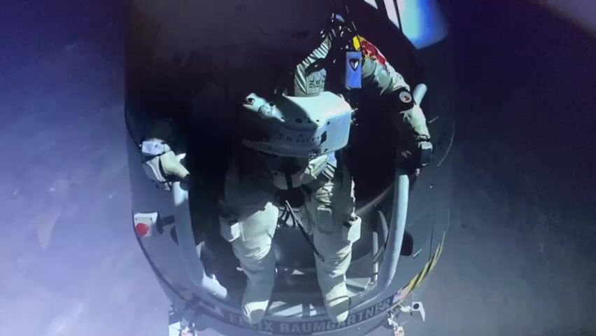 The video shows a man in a white spacesuit standing in an open hatchway, looking down. He is attached to a harness and appears to be preparing to jump. In the background, there is a man wearing headphones and speaking into a microphone, likely part of a mission control team. The environment appears to be a high-altitude or outer space setting, with a dark, blurry background. The man in the spacesuit is the primary focus of the scene, suggesting he is about to undertake a significant feat.