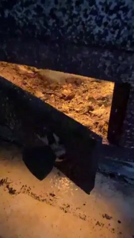 The video shows a close-up, low-angle shot of the interior of a fireplace or hearth. The main focus is on a triangular opening revealing a bed of glowing embers and ash. The texture of the embers is granular and appears to be emitting a warm, orange light. A dark, angled metal or ceramic piece, possibly part of the fireplace structure, protrudes into the foreground from the left, partially obscuring the opening. A small, dark, heart-shaped object, perhaps a piece of charcoal, is visible resting on the surface in front of this angled piece. The background is dark and textured, suggesting the rough, soot-covered interior of the fireplace. The lighting is dim, with the primary illumination coming from the glowing embers.