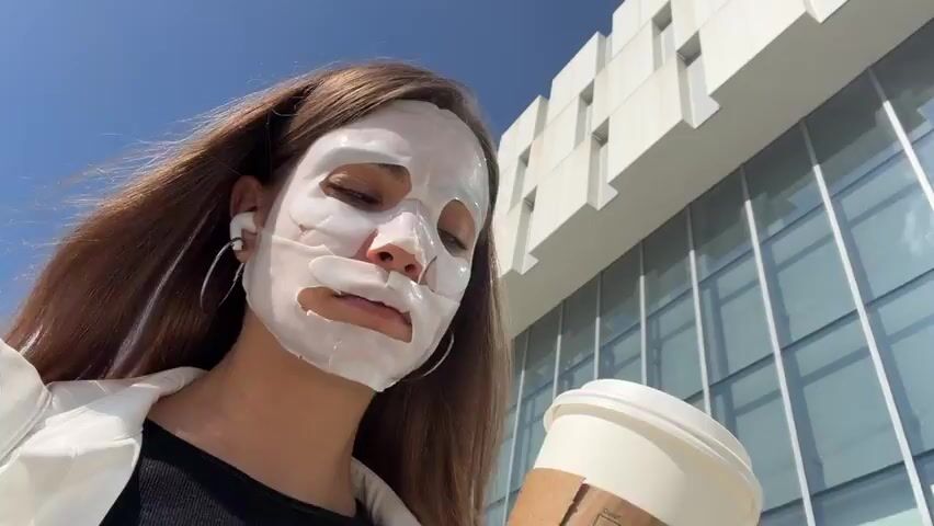 A woman wearing a sheet mask and earbuds holds a Starbucks coffee cup to her face and takes a sip. She is outdoors, with a building and a blue sky in the background.