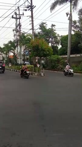 A street scene captured in daylight. A paved road with visible lane markings. Several motorcycles are present, with riders. One motorcycle is in the foreground, obscuring part of the view, with its rear wheel and fender clearly visible. To the left, another motorcycle is moving away from the camera. Further ahead, a light blue van is stationary at an intersection, with a person standing near it. A few trees and greenery are visible along the roadside, including palm trees. Utility poles with numerous wires are prominent in the background. The sky appears overcast.