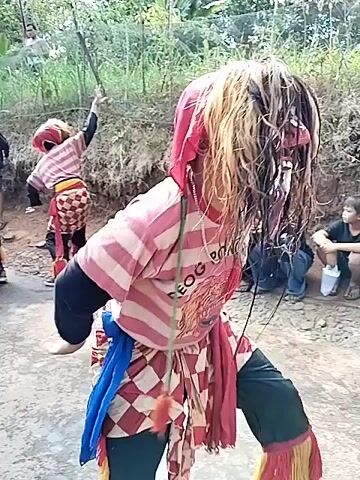 The video captures a traditional performance in an outdoor setting, possibly a rural area with trees and a dirt path. A performer in a striking costume, featuring a red and white striped shirt, a red head covering with long, wild blonde and brown hair, and a patterned skirt with a blue sash, is in the foreground. The performer's face is partially obscured by the hair, but appears to be wearing a mask. They are in a dynamic pose, with one arm extended upwards and the other bent. In the background, a group of spectators, including men, women, and children, are watching the performance. Some individuals are seated, while others are standing. Another performer, also in a traditional costume, is visible further in the background, near a body of water or muddy area. The overall atmosphere suggests a cultural event or celebration.