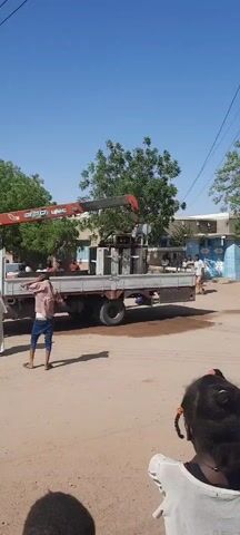 Here is a concise, forensic description of the video:

**Setting:** The video appears to be shot outdoors in a residential or commercial area with buildings and trees in the background. The ground is a dusty, unpaved surface. The sky is clear and blue.

**Objects:**
*   A flatbed truck is present, with a red hydraulic crane labeled "UNIC 330" mounted on it.
*   A large, metal electrical transformer unit sits on the truck's flatbed.
*   Electrical poles and wires are visible overhead.

**People:**
*   Several people are visible in the video.
*   One man wears a traditional white robe.
*   Another man wears a pink shirt and blue jeans.
*   Several people are standing near or behind the truck.

**Actions/Interactions:**
*   The crane is in operation, presumably lifting or securing the electrical transformer.
*   The man in the pink shirt walks in the foreground, partially obscuring the view.
*   The man in the white robe stands near the truck.
*   People are generally observing the operation.

**Environment:**
*   The overall environment seems to be a daytime scene in a warm climate, possibly in an urban area of a developing country.