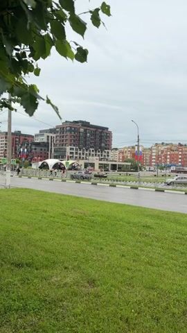 This is a video clip of a street scene in Obninsk, Russia, on an overcast day. In the foreground, lush green grass lines the side of a wet asphalt road. Across the road, a modern cityscape unfolds with several multi-story buildings. The architecture features a mix of styles, with some buildings displaying reddish-brown brickwork and others having lighter-colored facades. Flags, appearing to be Russian flags, are visible on poles in front of some buildings, suggesting a public space or event.

Several cars are driving on the road, indicating moderate traffic. Pedestrians can be seen walking on a sidewalk, some under umbrellas, suggesting it might be raining or has recently rained. A white tent-like structure is visible in front of one of the buildings. The sky is predominantly gray and cloudy, contributing to a subdued atmosphere. The overall impression is of a typical urban environment on a cloudy day. No text is clearly legible in the video.