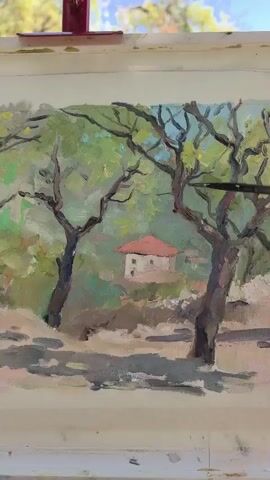 A close-up video shows an artist painting a landscape outdoors. The painting depicts two large, gnarled trees in the foreground with a small, white building with a red roof nestled in the background. The artist's hand and paintbrush are visible as they add details to the painting. The background of the video reveals the actual landscape the artist is painting, featuring more trees, sunlight, and a blue sky. The painting is on an easel, suggesting it's an plein air artwork.