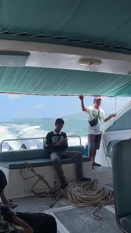 The video shows the interior of a boat moving through the water. In the foreground, two people are visible. On the left, a young man in a black long-sleeved shirt and gray pants is sitting on a bench and looking at his phone. In the middle of the frame, a coil of thick rope lies on the deck. On the right, a man in a white t-shirt, gray shorts, and a baseball cap stands and holds onto a railing. He is looking forward. The boat has two outboard motors visible in the back. The water behind the boat is turbulent and creates a wake. In the distance, green islands are visible under a blue sky with scattered clouds. The boat appears to be moving at a moderate speed.