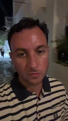 The video shows a close-up of a man with dark hair and a light complexion wearing a striped polo shirt. He appears to be outdoors at night, walking down a path lined with buildings and vegetation. The man is speaking to the camera and occasionally gestures with his hands. The environment suggests a warm climate, possibly a tourist destination. The lighting is dim, with artificial light sources illuminating the scene.