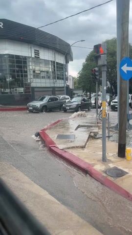 The video shows a flooded street corner in an urban environment. There are several vehicles navigating through the water, including a gray SUV and a dark gray pickup truck, which are both partially submerged. The water level reaches up to the bottom of the cars. The vehicles proceed cautiously through the intersection. A person in black clothing is seen walking on the sidewalk, crossing the street despite the flooding. A red traffic light is visible on a pole. The building on the corner has a modern design with large glass windows and the word "NOVAR" on the facade. The sky is overcast. The sidewalks have red edges, and the water is flowing over the curbs and into storm drains. A parked motorcycle is also seen by the traffic light. The scene is filmed from inside another vehicle.