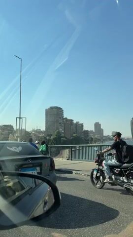 The video shows a scene from a vehicle driving over a bridge. In the foreground, the side mirror of the vehicle is visible on the left, reflecting the car in front. The car in front has a license plate. A motorcycle with a rider wearing a backpack is in the lane ahead. To the right, the side of a white bus is visible, with people visible inside the windows. The bus has Arabic writing on the back and a license plate. The bridge has a railing separating the road from a body of water. In the background, a skyline of buildings is visible across the water, with a clear blue sky above. The video pans slightly to the right, focusing more on the buildings.