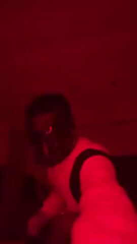 The video is shot in a room bathed in red light, making it difficult to discern specific details. A person with dark hair, wearing what appears to be a light-colored jacket with dark sleeves, is dancing. The person's face is obscured, and the movements are energetic and somewhat jerky. The background is blurry, but there seem to be furniture or equipment present. The room is relatively small and contains a few ceiling lights.