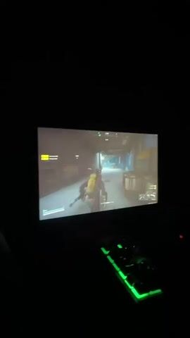 The video displays a first-person perspective of someone playing a video game in a dark room. The game appears to be a shooter or action-adventure, with the player character seen from behind, holding a weapon, moving through an indoor environment. A green-lit keyboard is visible in the foreground, suggesting it's being used for gameplay. The screen shows in-game menus and graphics, including a character model and a virtual environment. The overall setting is dimly lit, with the primary light source emanating from the monitor.