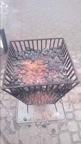 The video shows a square metal basket filled with burning coal. The basket is elevated on four legs above a light-colored rectangular tray. The ground beneath the tray is paved with gray bricks. The coal burns brightly in the center, emitting orange and yellow flames. Some embers can be seen among the coal pieces. A dark object is visible on the left, and the overall setting appears to be outdoors.