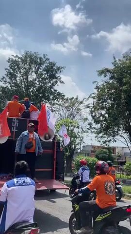 The video captures a dynamic procession or rally taking place on a sunny day in Tebel, Indonesia.

The primary subjects are a large group of people, predominantly men, involved in what appears to be an organized motorcade. Many participants wear matching orange t-shirts, suggesting a unified affiliation.

A red flatbed truck, functioning as a mobile stage and sound system, leads or accompanies the procession. Large speakers are mounted on its back. On the truck, a man in a light blue denim jacket, orange t-shirt, and cap stands prominently, seemingly addressing the crowd or leading chants, while other individuals in orange shirts are also visible on the truck bed. The side of the truck features a painted portrait of a smiling man.

Numerous white flags with a distinct blue and red logo are displayed. These flags prominently feature the acronym "FSPMI" (likely Federasi Serikat Pekerja Metal Indonesia – Federation of Indonesian Metal Workers' Union, given other visible text), along with "SERIKAT PEKERJA" (Worker's Union) and "KSPMI" on some. A man in the foreground wears a white and blue shirt with "FSPMI SPDT SIDOARJO" printed on it. Another orange shirt bears the text "AYDAY."

Behind the truck, a long line of motorcyclists, mostly on scooters or mopeds, follows. Many riders also wear the orange t-shirts, and some have the FSPMI flags attached to their motorcycles or held aloft.

The scene is set on a wide road lined with mature green trees, under a bright sky with scattered white clouds, indicating good weather. The organized attire, flags, and sound system suggest a planned demonstration, protest, or rally by a workers' union. The atmosphere appears lively and determined.