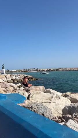 The video opens with a wide shot of a rocky shoreline on a clear, sunny day. The camera pans slightly to the left, revealing two people sitting on large rocks near the water. The person in the foreground is wearing a dark swimsuit and facing away from the camera, looking out at the ocean. Behind them, a second person is partially visible, also sitting on the rocks. The water is a vibrant blue-green, and a small white boat is visible in the distance. The background shows a breakwater or pier with more rocks and some distant buildings and palm trees. The camera then zooms in on the back of the head of the person in the foreground, showing their dark hair tied back in a low ponytail. They are wearing a dark, distressed t-shirt with holes along the shoulders. The video then returns to a wider shot of the same scene, with the two people still sitting on the rocks. The perspective shifts slightly, showing more of the blue-painted structure in the foreground. The scene is peaceful and depicts a relaxing day by the sea.