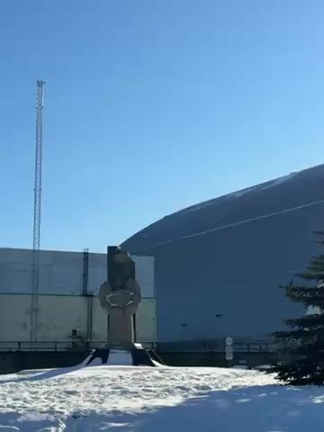 The video shows a wide shot of the Chernobyl Nuclear Power Plant sarcophagus in winter. In the foreground, there is a snow-covered ground with a monument and a few evergreen trees. In the background, the massive sarcophagus dominates the scene under a clear blue sky. The sun is visible behind the trees. A tall communications tower stands to the left. The camera pans slightly to the right. The overall scene is stark and cold.