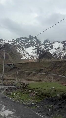 The camera pans slightly upward, revealing a mountain landscape beneath a cloudy sky. In the background, snow-capped mountains rise prominently, partially obscured by the overcast weather. The middle ground features terraced hills, with a road winding across them. A small, red bridge spans a stream in the foreground. A telephone pole stands on the left, with a power line cutting across the sky. The immediate foreground consists of a rough, rocky area with patches of green vegetation.  A gondola lift appears to be running on the ridge between the foreground and the mountain.