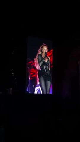 The video shows a concert from the perspective of an audience member. Initially, a screen displays a female singer in a black outfit, holding a microphone and performing. As the video progresses, the view shifts to the main stage, featuring a large circular logo lit in red, and a crowd of people facing the stage. The environment appears to be dark, with stage lighting illuminating the performers and the crowd.