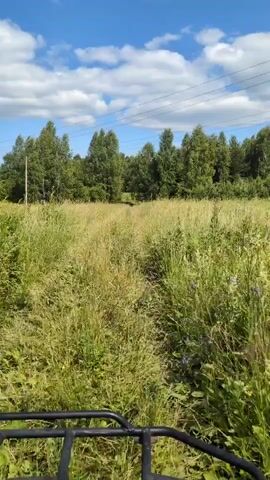 The video shows a first-person perspective of someone riding an ATV through a field of tall grass. The ATV is black and the handlebars are visible in the foreground. The field is wide and open with a dirt path leading through it. On either side of the path, the grass is tall and green, with some wildflowers interspersed. Beyond the field, there is a dense line of trees, likely a forest. Above the trees, a bright blue sky is visible with scattered white, fluffy clouds. Power lines are strung across the sky, supported by utility poles which are visible to the left. The overall impression is a sunny day in a rural, natural setting. The time of day appears to be daytime, likely midday given the brightness of the sky. The weather appears to be clear and pleasant, with a blue sky and clouds, suggesting it's a good day for outdoor activities. The location is identified as Velikovo, Russia. There is no visible text in the video. The scene evokes a sense of freedom, adventure, and enjoyment of nature.