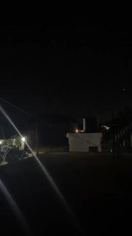 A short, dark video shows several houses illuminated by outdoor lights at night. There are some trees and power lines visible. The lights are bright and cause some lens flare in the video. The sky is black.