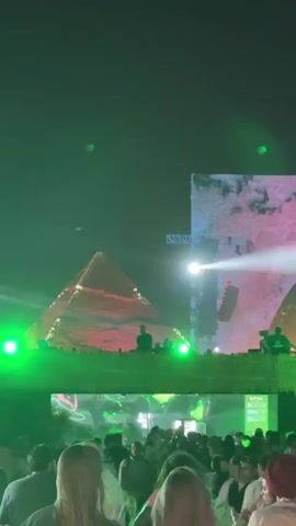 The video shows a music festival with a large stage and a crowd of people. The stage is decorated with a pyramid and an arch, both covered in pink and red designs. There are green lights illuminating the stage and the crowd. Two figures appear to be performing on stage. The crowd is mostly composed of young people who are standing close together and facing the stage. Some people are walking around, while others are dancing. The overall atmosphere is energetic and festive. The background is dark, suggesting the event is taking place at night.