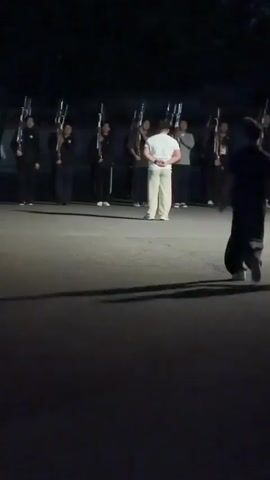 At night, in a large parking lot or open area, a group of people stand in a line holding what appear to be rifles with bayonets. A person in light-colored pants and a white shirt stands facing them. Two people are walking towards the group in the center. There are parked cars on the left side of the frame, and trees are visible in the background. Another group of people can be seen on the right, and a vehicle is parked further back in the scene. Overhead lighting illuminates the area.