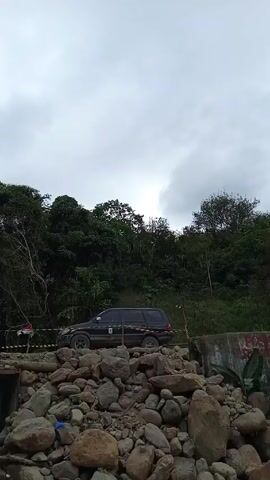The scene features a dark-colored SUV positioned atop a makeshift bridge constructed from large rocks and a concrete culvert. The vehicle appears to be slightly dirtied, suggesting it has been driven off-road. The landscape behind the SUV is dominated by lush, green trees and foliage, indicating a natural or forested area. The sky above is overcast with a layer of grey clouds, creating a diffused lighting condition. To the left, a person is observed holding a smartphone, seemingly capturing an image or video of the vehicle on the bridge. Red and white ribbons are draped across the rocks near the car. Water flows under the bridge.