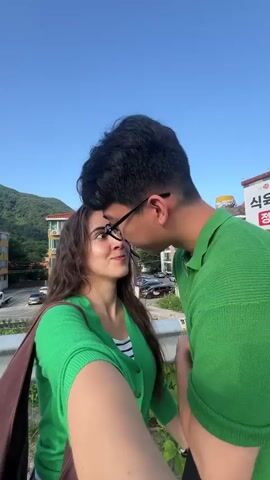 A young couple, a male and a female, are seen in a close-up shot, appearing to be kissing. The male, with dark hair and glasses, is positioned behind the female, with his head tilted down towards her. The female, with long brown hair, is looking up at him. Both are wearing green clothing. The background shows a clear blue sky, some buildings, and foliage, suggesting an outdoor setting. The video seems to be a selfie taken by the female, as her arm holding the camera is visible in the foreground. The overall atmosphere is intimate and romantic.
