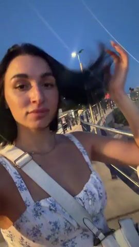 A young woman with dark hair and brown eyes, wearing a white floral dress and a white crossbody bag, is standing outdoors at dusk. She is looking at the camera, touching her hair, and smiling. In the background, there is a body of water, a railing, a street light, and some people. The woman gives a thumbs-up to the camera.
