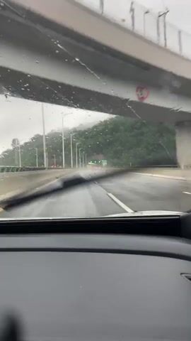 The video shows a view from inside a car driving on a wet highway. The windshield has raindrops and streaks, and the dashboard is visible in the foreground. The highway has multiple lanes, and the road surface is dark and appears wet. There are streetlights and guardrails along the side of the road. On the right, a lush green, tree-covered mountain slopes upward. Ahead, the highway leads towards a tunnel entrance, marked by green lights. The sky is overcast and gray.