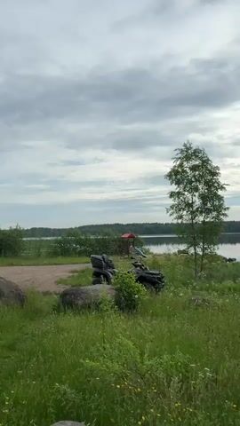 A person films a natural landscape featuring a lake and grassland under a cloudy sky. The video pans across the scene, showing a tall tree with bare branches, a small children's playground with a red slide, two ATVs, a wooden swing set, and a dense forest line in the distance. The lake's surface reflects the sky. The grass is lush and green, with scattered white and yellow wildflowers.