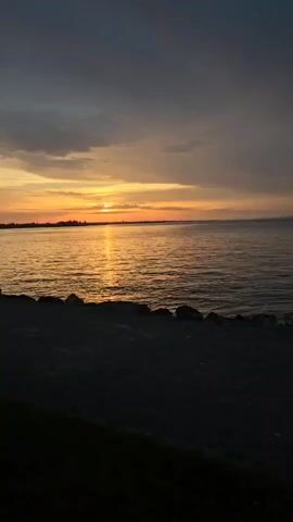 A video captures a sunset over a body of water. The sun, a bright yellow orb, is low on the horizon, casting a shimmering golden path across the waves. The sky is a mix of dark gray clouds and streaks of orange and yellow light. In the foreground, the dark silhouette of a rocky shore leads to the water. A person is partially visible on the left side of the frame, standing near the shore. The overall atmosphere is tranquil and serene.