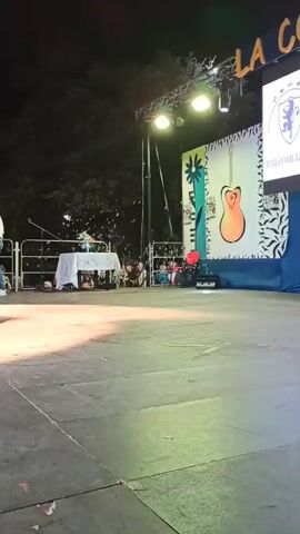 A man in a blue shirt and white pants stands on a stage, holding a microphone to his mouth and singing. He is wearing glasses and has gray hair. He is illuminated by a spotlight. Behind him, a backdrop features a stylized drawing of a guitar. In the background, there is a crowd of people seated and standing, watching the performance. There are also stage lights, speakers, and other equipment visible on the stage and in the surrounding area. The scene appears to be an outdoor event at night, with trees visible in the background.