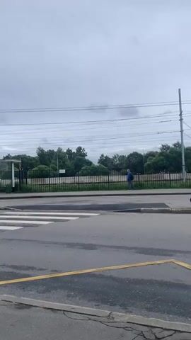 The video shows a train station on a cloudy day. In the foreground, there is a crosswalk with yellow lines on the asphalt road. A black metal fence runs along the train tracks, separating them from the road. Beyond the fence, there are trees and a few buildings. A blue train with white lettering that reads "РЖД" (Russian Railways) is visible on the tracks. A person wearing a blue jacket is standing near the fence, looking towards the train. The sky is overcast.