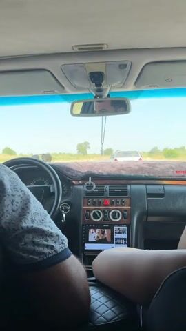 The video shows the interior of a car from the back seat, looking forward. The driver, a man with short dark hair, is visible on the left. A woman with long red hair is partially visible on the right. The dashboard of the car, including the steering wheel, radio, and air vents, is in the foreground. Outside the car, a road with other vehicles can be seen under a clear sky. The car appears to be moving.
