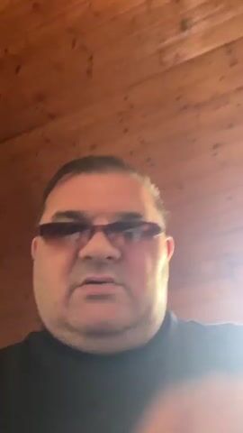 The video shows a close-up of a man wearing sunglasses and a black t-shirt against a wood-paneled background. The man is gesturing with his right hand, with his thumb and index finger held close together. The camera is positioned at a low angle, looking up at the man. The lighting is warm, with some glare on the man's sunglasses.