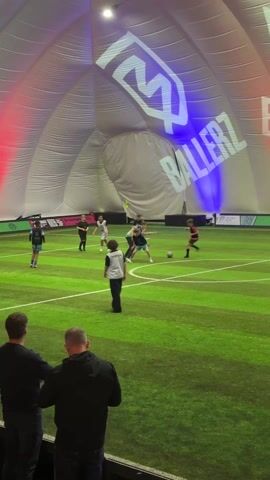 The video shows a group of young people playing soccer in an indoor facility called Ballerz. The players are wearing different colored uniforms, and they are moving around the green field, passing and kicking the ball. Two men are standing in the foreground, watching the game. The facility has an inflatable roof with the "Ballerz" logo projected on it in blue and red lights.