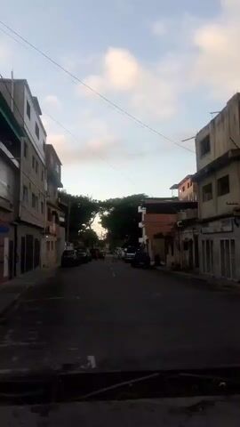 The video shows a street scene in a city. The street is lined with multi-story buildings on both sides. The buildings appear to be made of brick and concrete. There are cars parked along the street. Some people can be seen walking in the distance. The sky is visible above the buildings, with some clouds present. The overall impression is that of a residential or mixed-use urban environment. The lighting suggests it is either early morning or late afternoon.