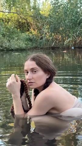 A woman with brown hair in two braids is in a body of water, likely a river or lake. She is submerged from the chest up and is wearing a sheer, light-colored garment. She is holding a dark-colored rope or braid of hair in her hands and appears to be bringing it to her mouth. The background shows dense green foliage and trees, with the water reflecting the surrounding environment. The lighting suggests it is daytime.