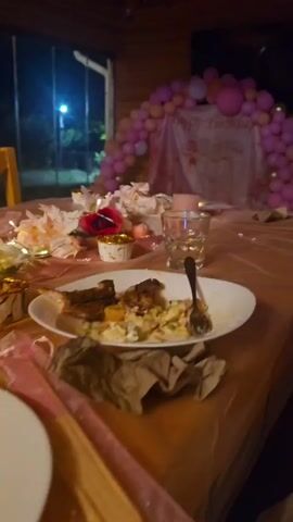 The video pans across a table laden with the remnants of a meal. Plates are strewn about, some with leftover food, others scraped clean. Forks and other utensils lie scattered amongst crumpled napkins. A half-eaten chocolate cake sits on one plate, its slices unevenly cut. Several glasses are visible, one containing a yellow beverage, others empty. Decorative elements, including pink balloons forming an arch in the background and scattered floral pieces, suggest a celebration, possibly a birthday party. The setting appears to be outdoors or in a covered patio area, with a glimpse of lights and trees in the dark background. The overall impression is of a post-event scene, where a gathering has concluded, leaving behind the mess of a shared meal.
